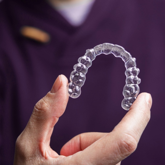 Mão segurando um alinhador dental transparente em foco, com fundo desfocado em tom roxo, destacando o produto.