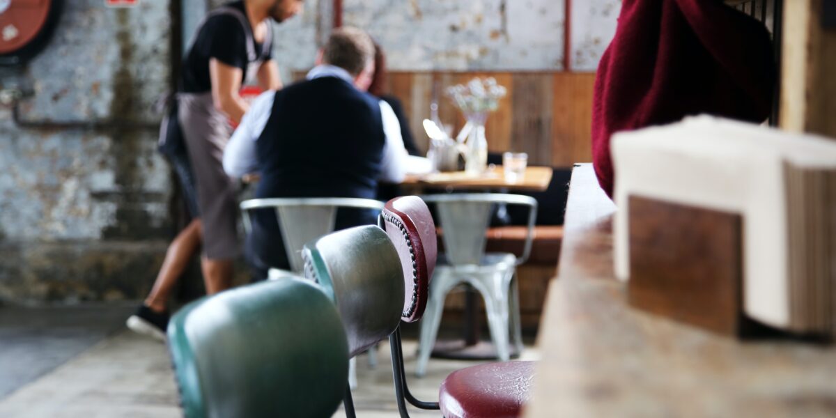 Cadeiras em primeiro plano e pessoas sendo atendidas em mesa de restaurante ao fundo.