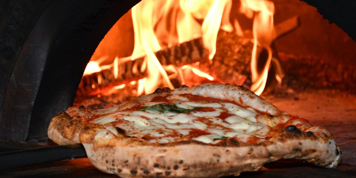 Pizza recém assada sendo retirada de forno a lenha.