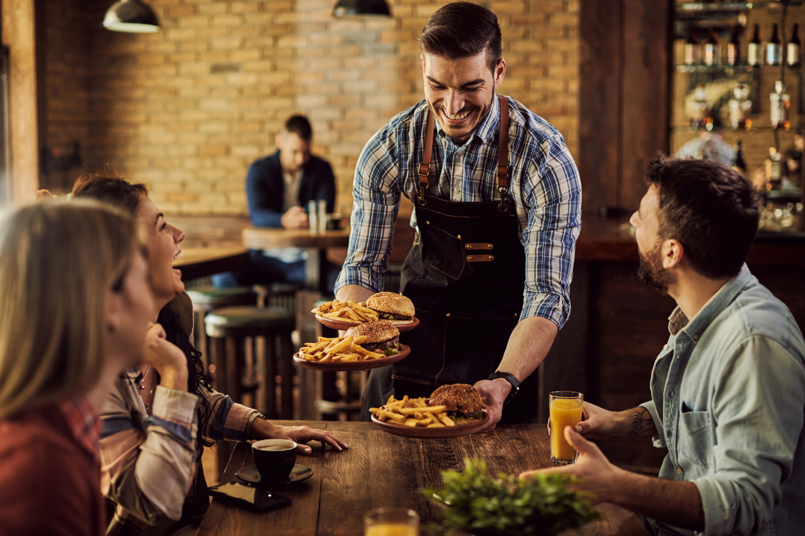 Atendente sorridente anota o pedido de clientes em um restaurante iluminado e aconchegante, com mesas ocupadas ao fundo.