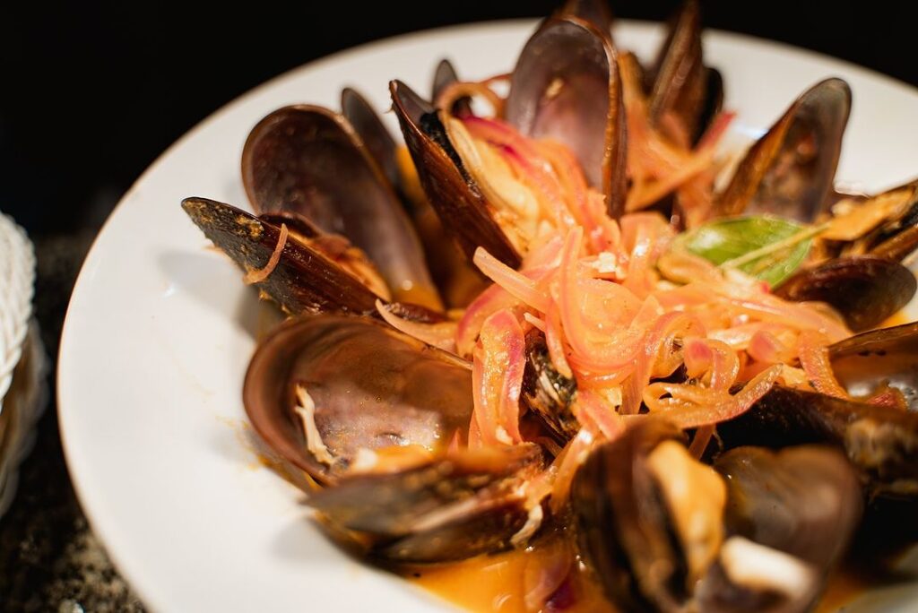 Prato de mexilhões ao molho, servidos em prato branco com cebola e ervas.