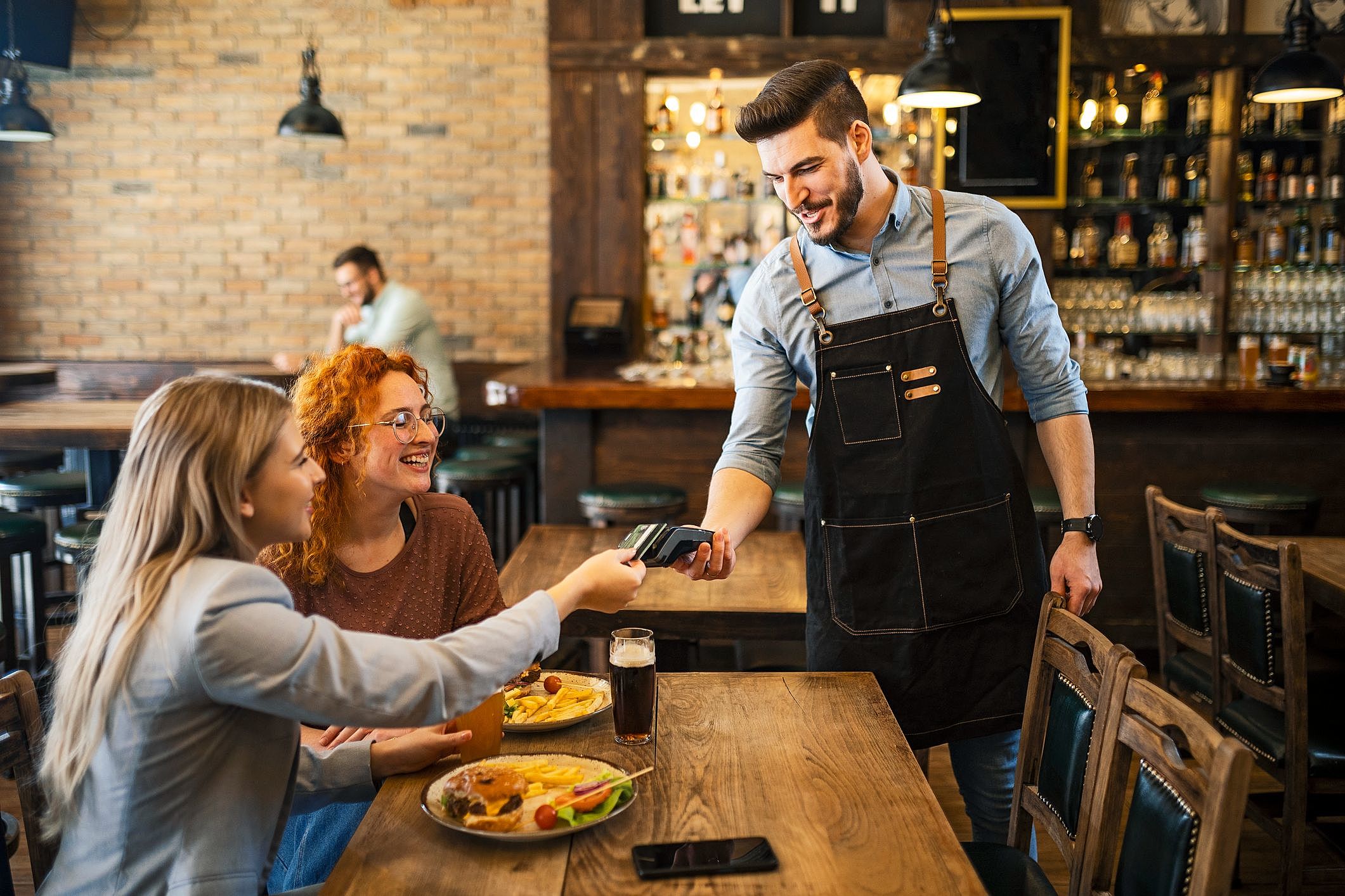 Garçom entrega a conta em máquina de pagamento enquanto clientes estão sentados à mesa em restaurante.