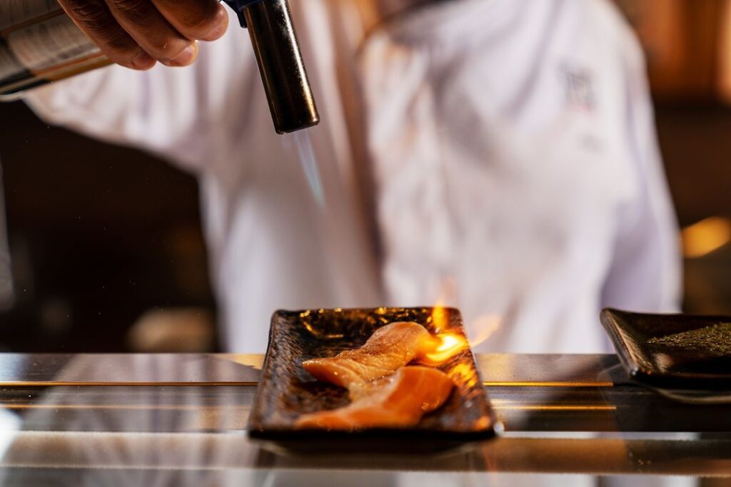 Chef finaliza peixe com maçarico sobre um prato em bancada de cozinha profissional.